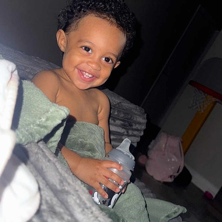 Espoir joined the competition — help win amazing prizes! toddler, child, smiling, baby_bottle, towel, curly_hair, indoor, couch, blanket, toy, basketball_hoop, carpet, happy, person, home, furniture, cute, young_child, cozy, relaxed
