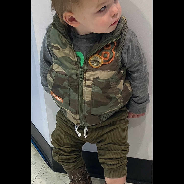 Creed is registered to the contest to win money with this photo: boy, camouflage_vest, candid, casual_clothing, child, cowboy_boots, curious, fashion, floor_tiles, footwear, gray_shirt, indoor, looking_away, olive_pants, portrait, short_hair, standing, toddler, white_wall, young_child