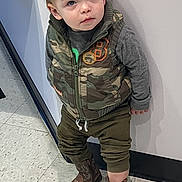 Creed is registered to the contest to win money with this photo: toddler, child, boy, standing, camouflage_vest, cowboy_boots, indoor, floor, wall, curious, clothing, young, person, portrait, casual, boots, pants, long_sleeve_shirt, hair, expression