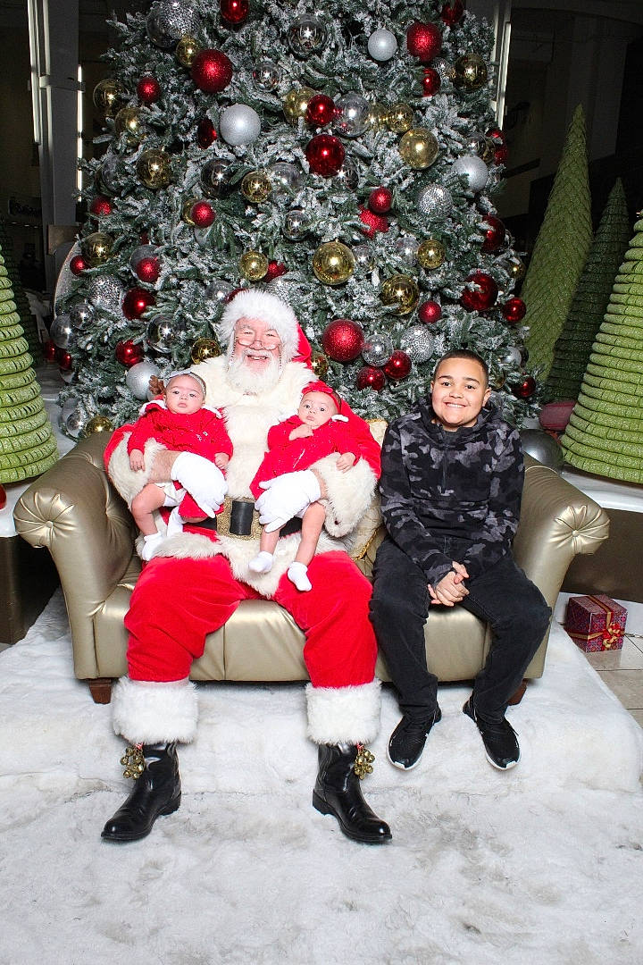 Alani is registered to the contest to win money with this photo: boot, christmas, christmas_decoration, christmas_eve, christmas_ornament, christmas_tree, conifer, event, fun, headwear, holiday, holiday_ornament, joy, lap, leg, person, plant, santa_claus, sitting, smile