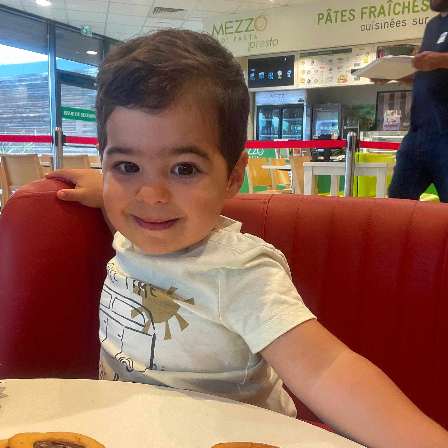 Tiago participe au concours pour gagner de l'argent avec cette photo : casual_dining, chair, child, cookie, curly_hair, food, happy, indoor, man, menu_board, person, red_bench, restaurant, smile, snack, table, toddler, tray, white_shirt, window