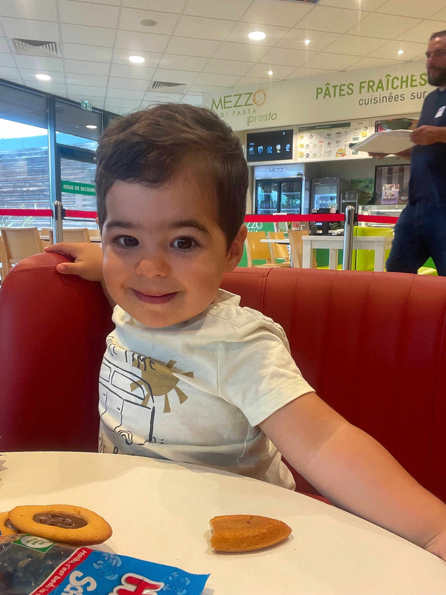 Tiago participe au concours pour gagner de l'argent avec cette photo : casual_dining, chair, child, cookie, curly_hair, food, happy, indoor, man, menu_board, person, red_bench, restaurant, smile, snack, table, toddler, tray, white_shirt, window