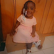 Aaliyah joined the competition — help win amazing prizes! child, toddler, girl, smile, pink_dress, white_sandals, beaded_hair, cardboard_box, wooden_wall, indoor, tile_floor, happy, sitting, cute, portrait, person, young_child, casual, home, joyful