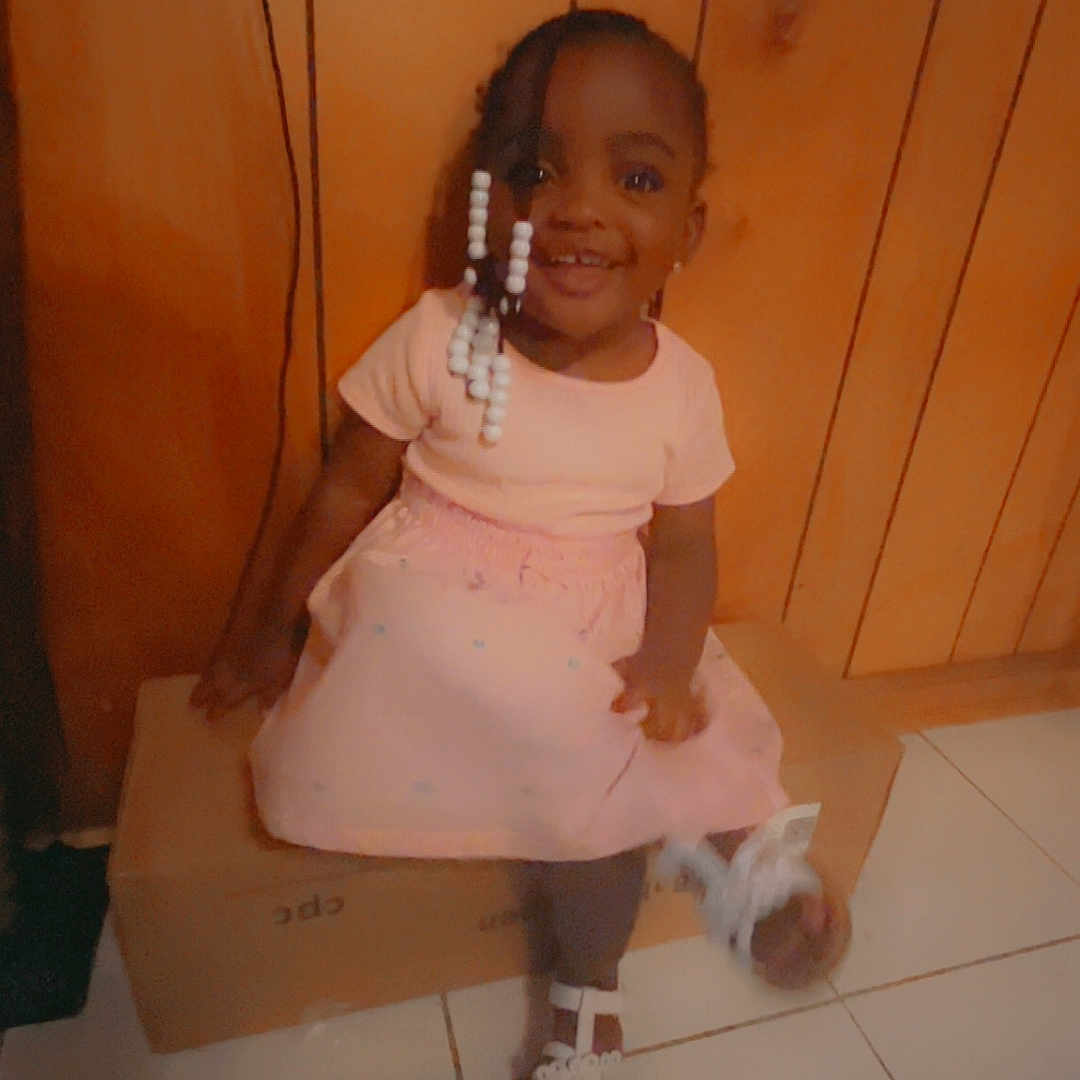 Aaliyah joined the competition — help win amazing prizes! child, toddler, girl, smile, pink_dress, white_sandals, beaded_hair, cardboard_box, wooden_wall, indoor, tile_floor, happy, sitting, cute, portrait, person, young_child, casual, home, joyful