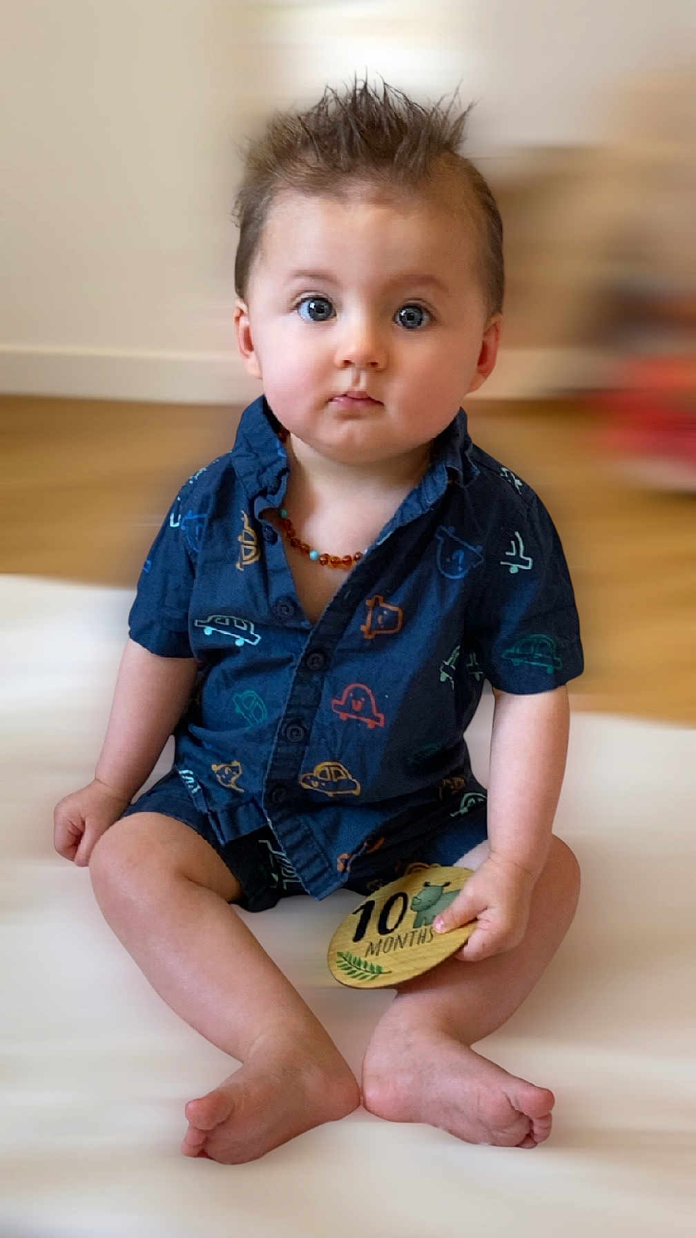Maylon a rejoint le concours — aidez-le/la à gagner de superbes lots ! baby, child, infant, sitting, blue_clothing, short_sleeves, car_pattern, wooden_plaque, milestone, indoor, floor, feet, hands, face, eyes, hair, necklace, expression, cute, portrait