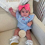 Hadley is registered to the contest to win money with this photo: baby, child, pink_bow, blue_dress, white_shoes, frilly_socks, armchair, plaque, milestone, indoor, smiling, happy, cute, portrait, infant, furniture, blanket, crib, raised_hand, person