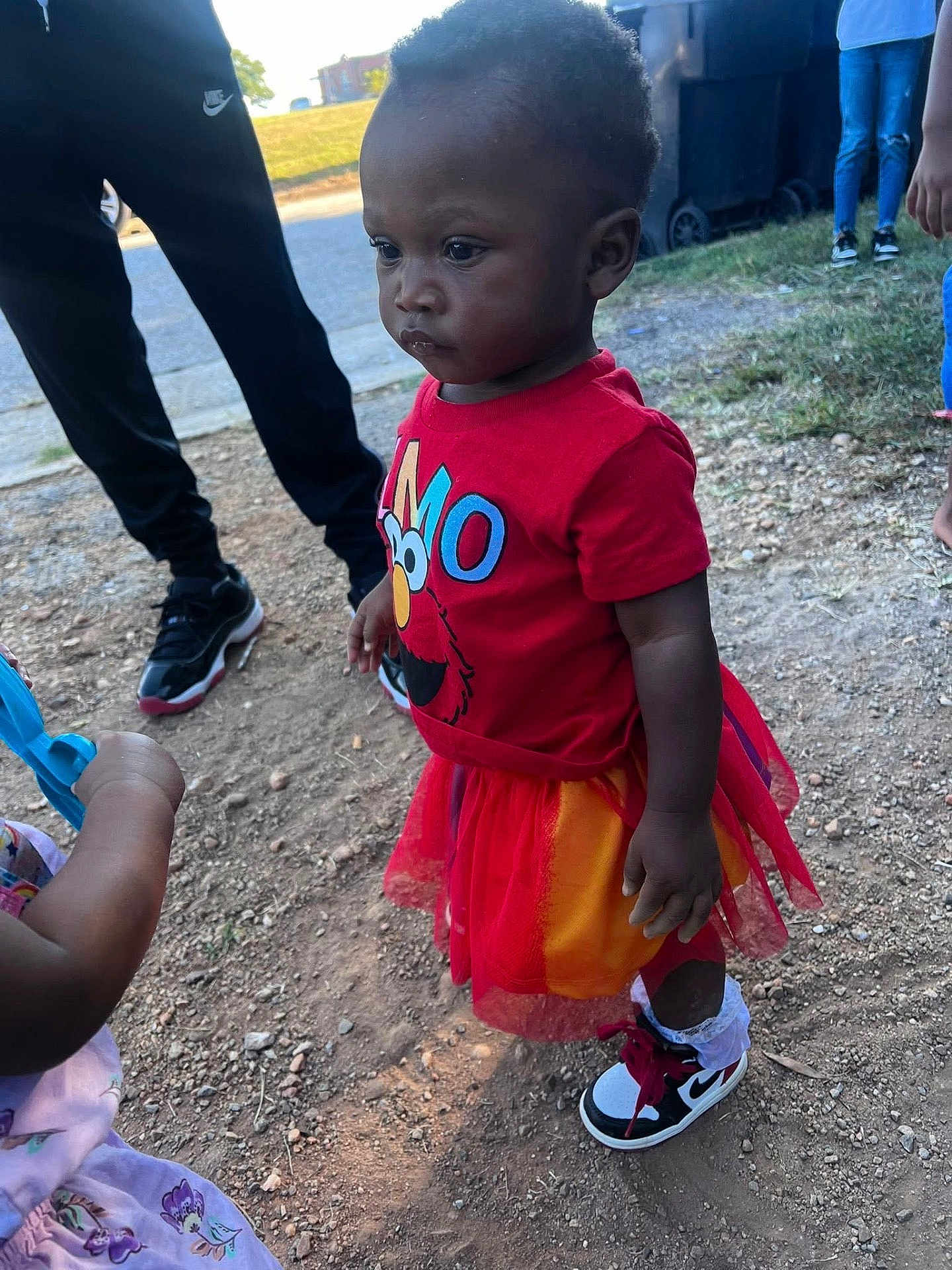 Mckenzie is registered to the contest to win money with this photo: toddler, child, red_shirt, elmo, tutu, outdoor, gravel, shoes, nike, socks, people, grass, sidewalk, casual_clothing, daylight, serious_expression, standing, toy, playing, group