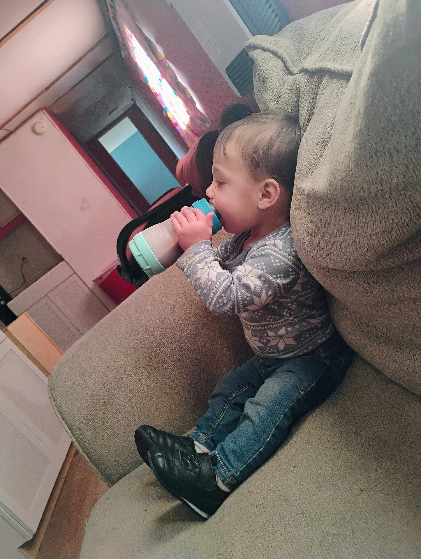 Nathan joined the competition — help win amazing prizes! toddler, child, bottle, sitting, couch, indoor, clothing, sweater, jeans, shoe, face, person, window, curtain, floor, furniture, room, light, baby, drink