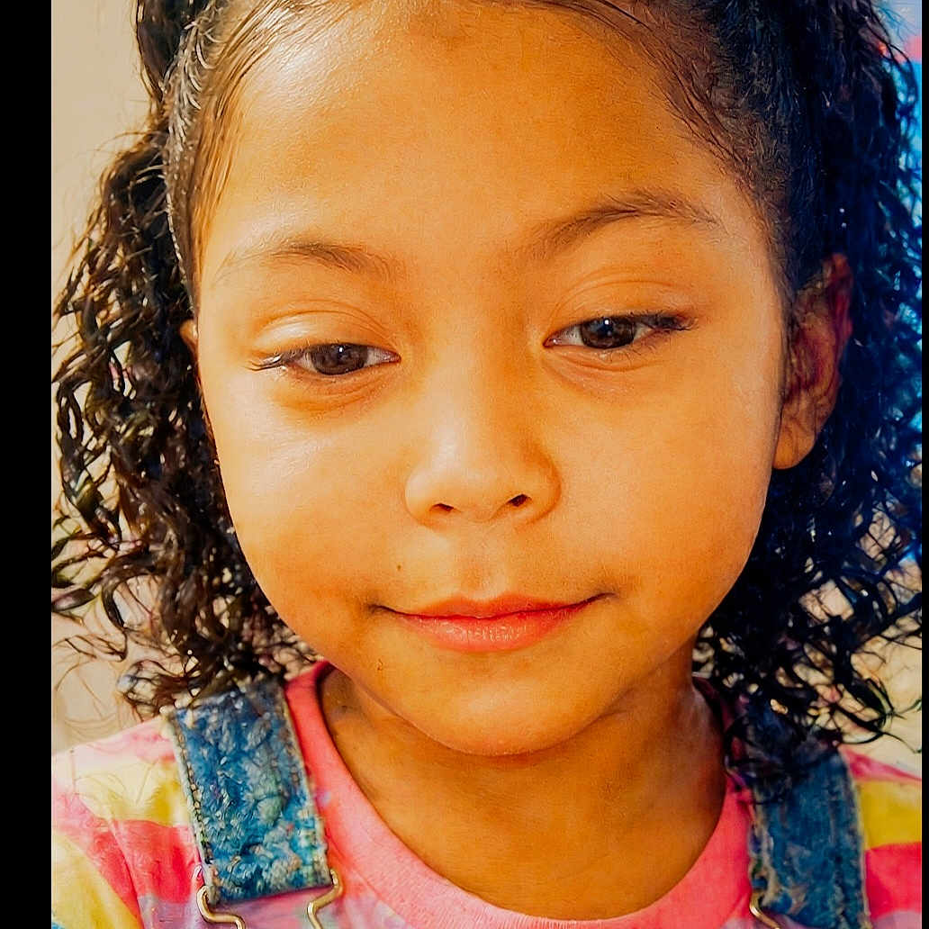 Athena is registered to the contest to win money with this photo: child, portrait, face, curly_hair, denim_overalls, colorful_clothing, smile, young_girl, casual_wear, indoor, close_up, cute, happy, person, eyelashes, skin, fashion, childhood, hair, expression