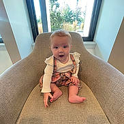 Charlotte is registered to the contest to win money with this photo: baby, child, armchair, plaid, romper, white_shirt, window, greenery, indoor, smiling, sitting, cozy, cute, infant, person, furniture, natural_light, happy, baby_clothes, room