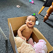 Ella is registered to the contest to win money with this photo: baby, cardboard_box, plush_toys, smile, indoor, carpet, happy, child, furniture, toy, candid, sitting, cute, person, floor, small_child, joyful, playful, home, necklace