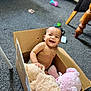 baby, cardboard_box, plush_toys, smile, indoor, carpet, happy, child, furniture, toy, candid, sitting, cute, person, floor, small_child, joyful, playful, home, necklace