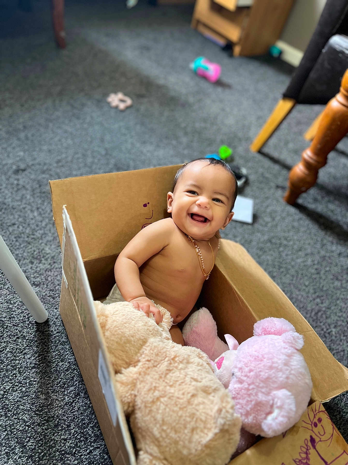Ella is registered to the contest to win money with this photo: baby, cardboard_box, plush_toys, smile, indoor, carpet, happy, child, furniture, toy, candid, sitting, cute, person, floor, small_child, joyful, playful, home, necklace