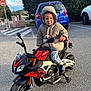 child, toddler, hoodie, jacket, toy_motorcycle, aprilia, training_wheels, cookie, smile, parking_lot, blue_car, pavement, outdoor, trees, hedge, hood_up, jeans, sneakers, handlebars, no_entry_sign