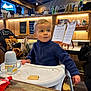 child, toddler, highchair, sippy_cup, cookie, cafe, wooden_interior, menu_clipboard, string_lights, barista, counter, coat, chair, table, snack, portrait, candId, window, cup, casual_clothing