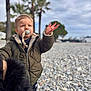 child, toddler, pacifier, jacket, beach, pebbles, palm_trees, cloudy_sky, raised_hand, outdoors, portrait, shallow_depth_of_field, coat, winter_clothing, serious_expression, standing, stones, blurred_background, tracksuit_pants, candid