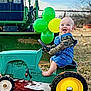 baby, child, toy_tractor, overalls, denim, camo_clothing, balloons, green, yellow, outdoor, grass, tractor, happy, smiling, barefoot, play, farm, vehicle, portrait, daytime