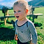 toddler, child, outdoor, grass, sunlight, blond_hair, striped_shirt, nature, picnic_table, blue_sky, greenery, casual_clothing, portrait, daylight, curious, young_child, expression, person, playful, background_blur