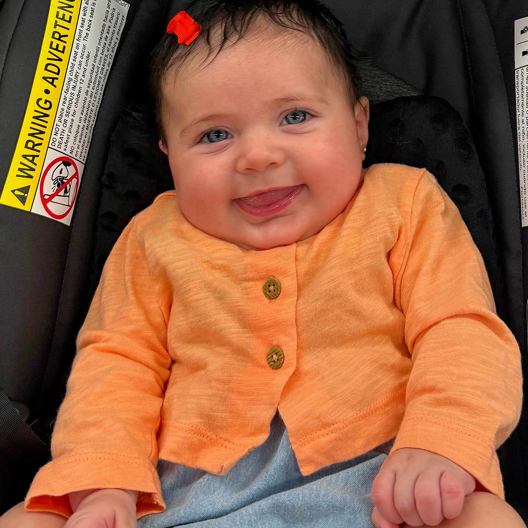 Manuela is registered to the contest to win money with this photo: baby, child, infant, smiling, tongue_out, orange_cardigan, hair_clip, car_seat, warning_label, cute, happy, portrait, sitting, clothing, face, hand, skin, eyes, person, indoors
