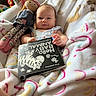 baby, blanket, book, child, colorful, cozy, doll, face, furniture, indoor, infant, person, plush_toy, reading, smile, soft_toy, text, toy, wearing_clothes, white_blanket