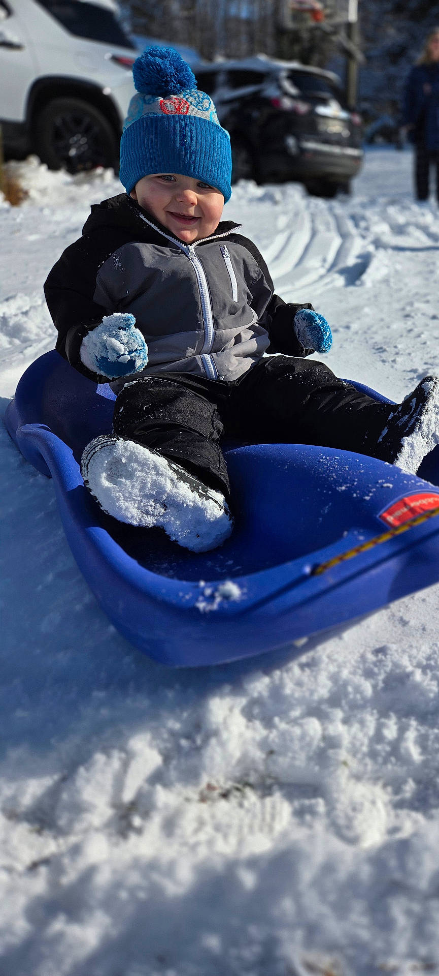 Ryder is registered to the contest to win money with this photo: blue_hat, child, cold_weather, daylight, frozen, happy, mittens, outdoor, parked_cars, playful, recreation, sled, smiling, snow, snow_boots, snowy_ground, toddler, winter, winter_clothing, winter_fun