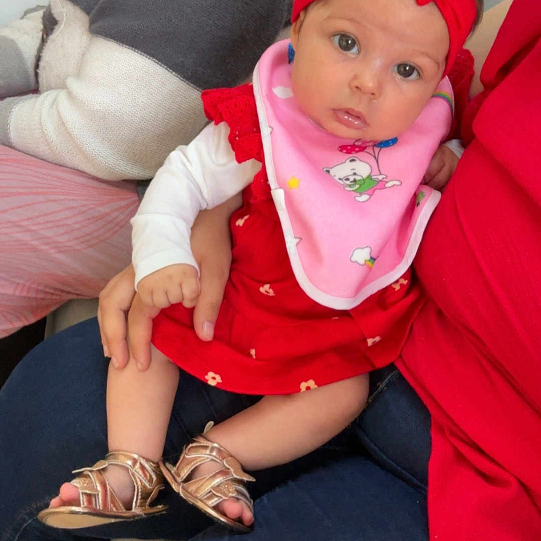 Elalïa a rejoint le concours — aidez-le/la à gagner de superbes lots ! adult, baby, child, clothing, curly_hair, face, family, footwear, hand, headband, indoor, infant, legs, looking, person, pink_bib, red_dress, sandals, seated, sitting