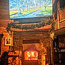 cat, fireplace, fire, scratching_post, wood_logs, television, indoor, cozy, rustic, living_room, stone_masonry, wooden_walls, decorations, garland, furniture, warm_lighting, cat_perched, home_interior, firewood, electronics