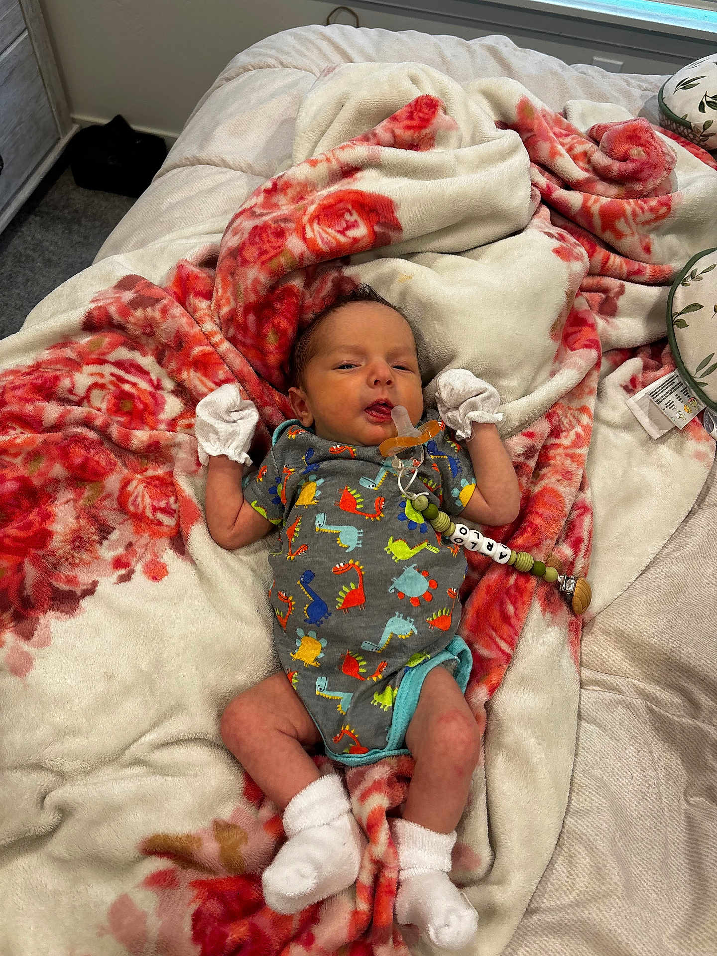 Arlo is registered to the contest to win money with this photo: baby, infant, blanket, floral_pattern, onesie, dinosaur_print, mittens, socks, pacifier, clip, bed, soft, cozy, indoor, person, newborn, cute, resting, comfort, sleepwear