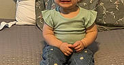 Penelope is registered to the contest to win money with this photo: toddler, child, smiling, sitting, barefoot, braids, denim, pants, green_top, cushion, pillow, indoor, carpet, happy, cute, portrait, young_child, casual_clothing, home, cozy