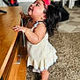 María is registered to the contest to win money with this photo: toddler, child, indoor, carpet, wooden_table, headband, flower, white_dress, shoes, standing, curious, happy, person, baby, furniture, hair, skin, expression, side_view, home