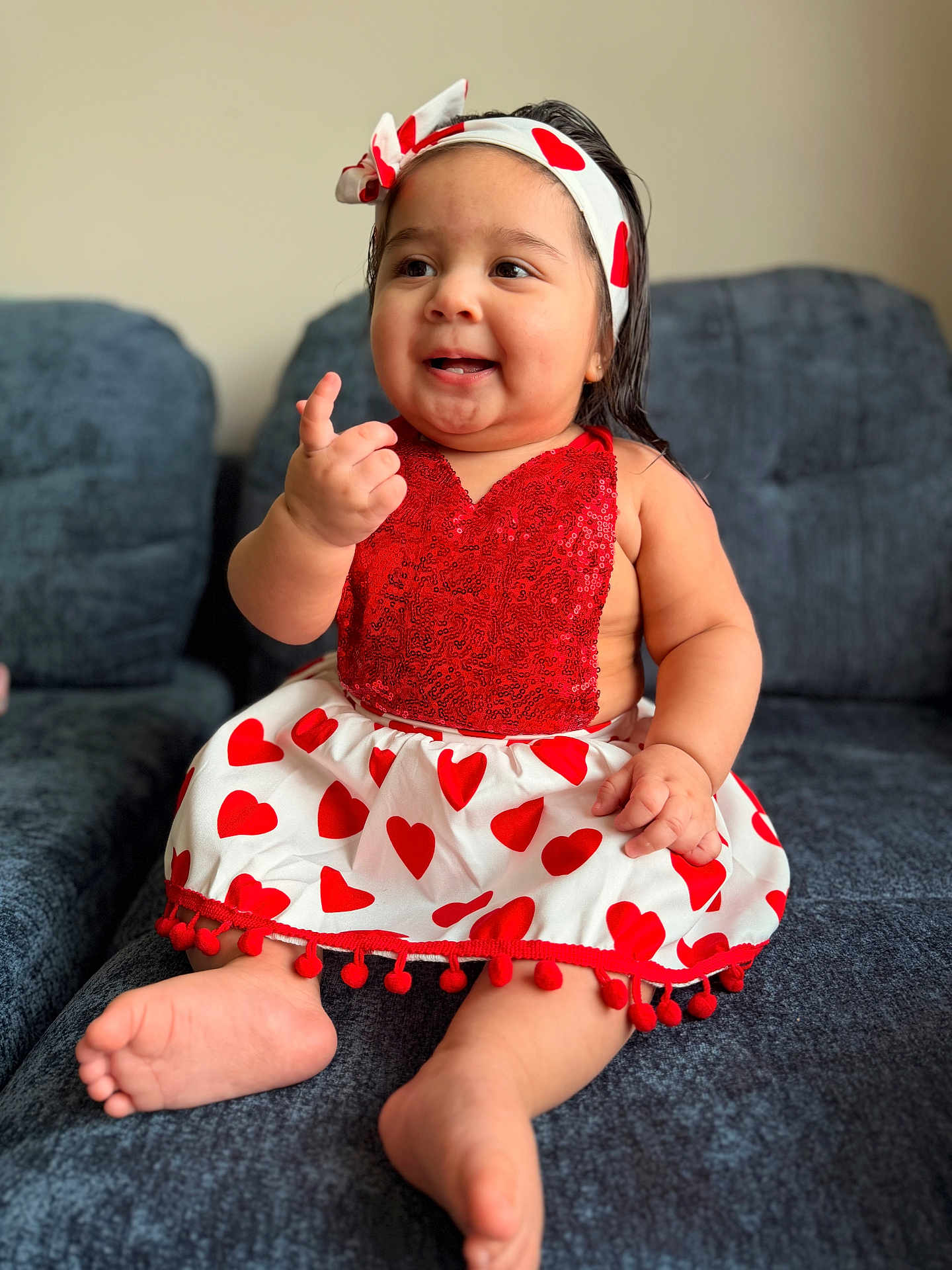 María is registered to the contest to win money with this photo: baby, child, smiling, sitting, dress, headband, heart_pattern, red, white, couch, indoor, cute, cheerful, happy, portrait, infant, person, furniture, skin, foot