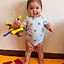 María is registered to the contest to win money with this photo: toddler, child, standing, plush_toy, smile, indoor, blanket, barefoot, onesie, cute, happy, baby, playful, toy, wall, floor, skin, person, young_child, expression