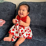 María is registered to the contest to win money with this photo: baby, child, smiling, happy, dress, red, white, heart_pattern, sitting, couch, indoor, cute, infant, portrait, clothing, person, foot, hand, joy, fun