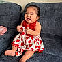 baby, child, smiling, happy, dress, red, white, heart_pattern, sitting, couch, indoor, cute, infant, portrait, clothing, person, foot, hand, joy, fun