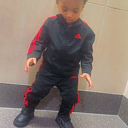 Azay is registered to the contest to win money with this photo: child, toddler, tracksuit, adidas, black_clothing, red_stripes, shoes, standing, step, tile_wall, indoor, curly_hair, looking_down, floor, young_child, casual_wear, pose, concentration, sporty, footwear