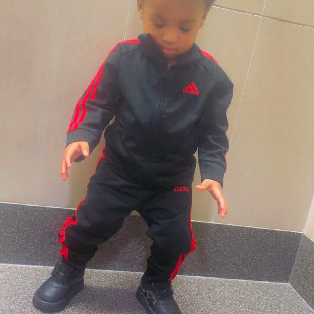 Azay is registered to the contest to win money with this photo: adidas, black_clothing, casual_wear, child, concentration, curly_hair, floor, footwear, indoor, looking_down, pose, red_stripes, shoes, sporty, standing, step, tile_wall, toddler, tracksuit, young_child