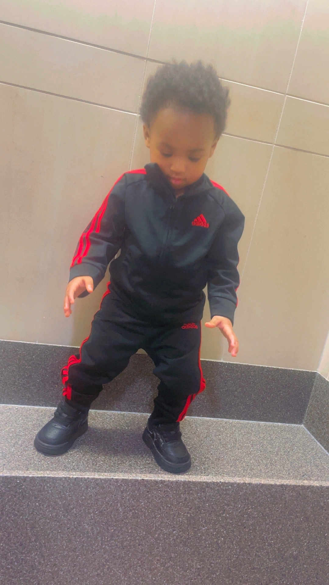 Azay is registered to the contest to win money with this photo: child, toddler, tracksuit, adidas, black_clothing, red_stripes, shoes, standing, step, tile_wall, indoor, curly_hair, looking_down, floor, young_child, casual_wear, pose, concentration, sporty, footwear