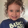 child, toddler, curly_hair, pigtails, smiling, blue_shirt, text_on_clothing, indoor, portrait, face, happy, person, casual_clothing, cute, young_child, head, eyes, skin, hair, expression