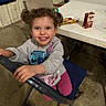 Novaleigh is registered to the contest to win money with this photo: toddler, child, smiling, indoor, folding_chair, table, food, drink, carton, pigtails, clothing, sweatshirt, pants, concrete_floor, chair, happy, person, portrait, casual, candid