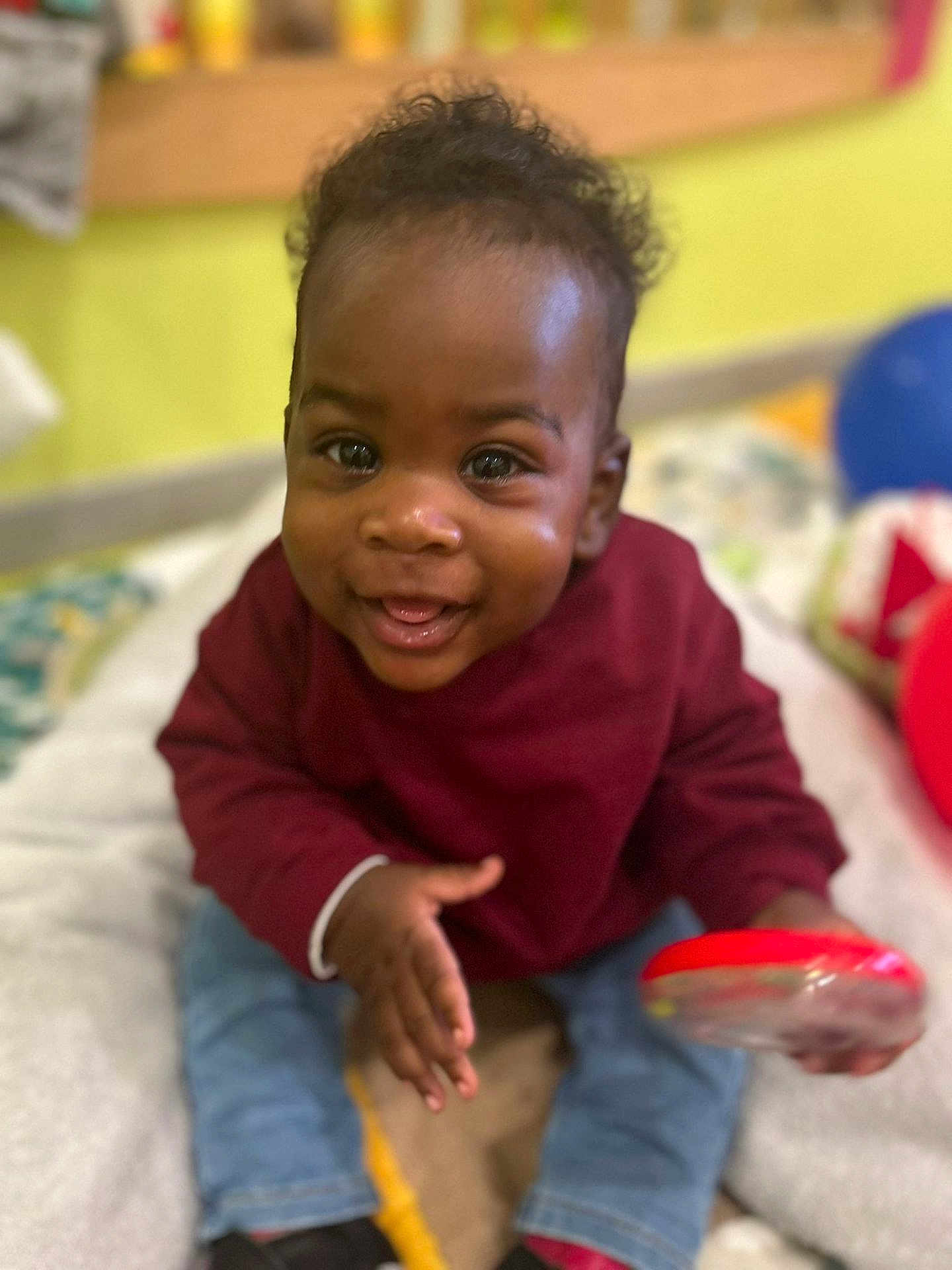 Ilétou a rejoint le concours — aidez-le/la à gagner de superbes lots ! baby, child, smiling, happy, indoor, toy, clothing, maroon_sweater, jeans, black_shoes, curly_hair, face, person, infant, cute, playful, sitting, bright, colorful, young