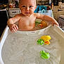 baby, bath, bathtub, rubber_duck, toy_frog, water, kitchen, child, wet_hair, playful, smiling, toddler, plastic_toys, sitting, indoor, cute, person, skin, face, expression
