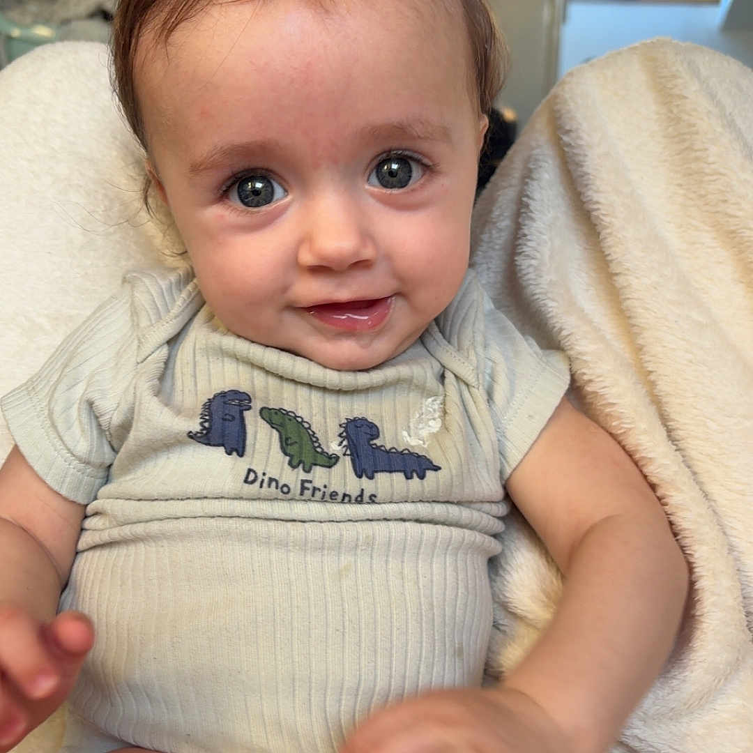 Aïden participe au concours pour gagner de l'argent avec cette photo : baby, infant, child, onesie, dinosaur_print, smiling, blue_eyes, soft_blanket, indoor, cozy, cute, playful, hand, legs, head, face, person, home, natural_light, portrait