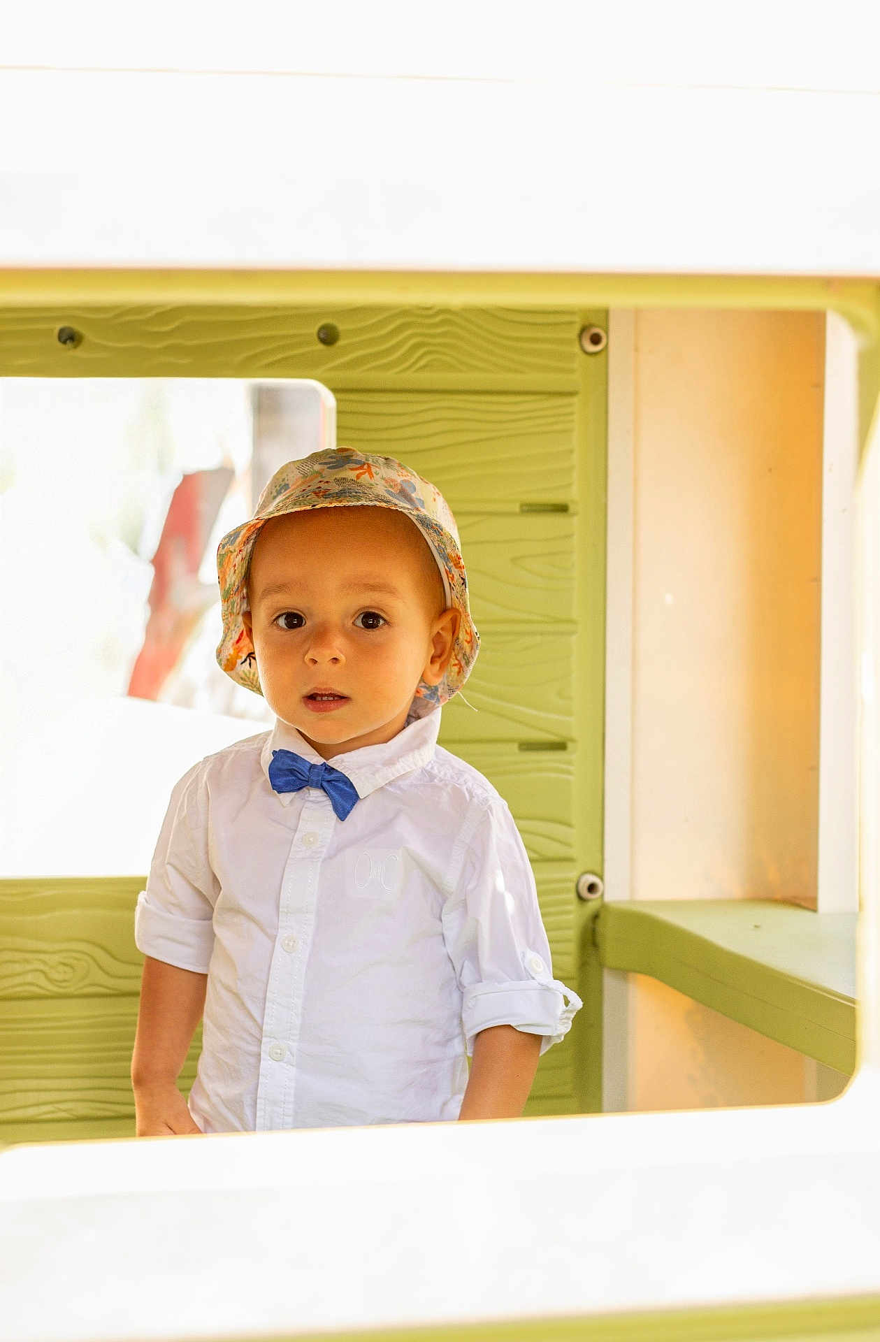 Lucien participe au concours pour gagner de l'argent avec cette photo : toddler, child, hat, bucket_hat, bow_tie, white_shirt, playhouse, green_wall, curious, portrait, indoor, young_child, casual_clothing, face, expression, person, standing, cute, fashion, light