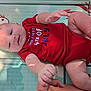 baby, infant, child, red_onesie, play_mat, hand, foot, skin, face, smile, light_shadow, text_on_clothing, celebration, holiday, cute, indoors, person, lying_down, young, toddler