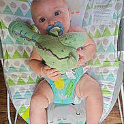 Benjamin is registered to the contest to win money with this photo: baby, blue_eyes, bouncer, child, chubby_legs, colorful, comfort, crocodile, cute, holding, indoor, infant, knitted_toy, pacifier, patterned_fabric, playful, seated, smiling, toy, wooden_floor