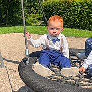 Eliano a rejoint le concours — aidez-le/la à gagner de superbes lots ! child, toddler, swing, playground, outdoor, rope_swing, grass, trees, sunlight, shadow, red_hair, blue_pants, white_shirt, bow_tie, suspenders, shoes, hand, person, gravel, nature