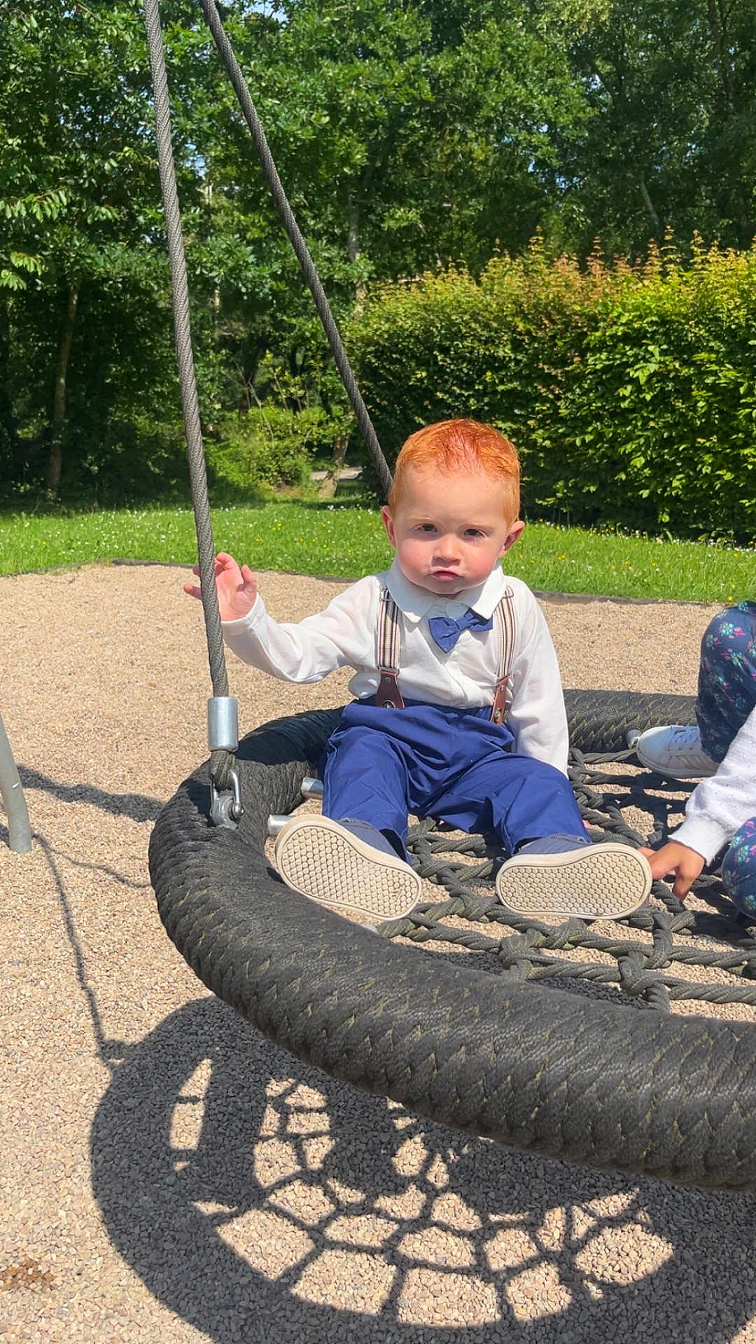 Eliano a rejoint le concours — aidez-le/la à gagner de superbes lots ! child, toddler, swing, playground, outdoor, rope_swing, grass, trees, sunlight, shadow, red_hair, blue_pants, white_shirt, bow_tie, suspenders, shoes, hand, person, gravel, nature