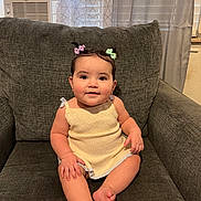 Tracey is registered to the contest to win money with this photo: baby, child, smiling, sitting, couch, gray_couch, hair_bows, indoors, window_air_conditioner, curtains, cute, portrait, infant, happy, skin, clothing, furniture, home, person, relaxed