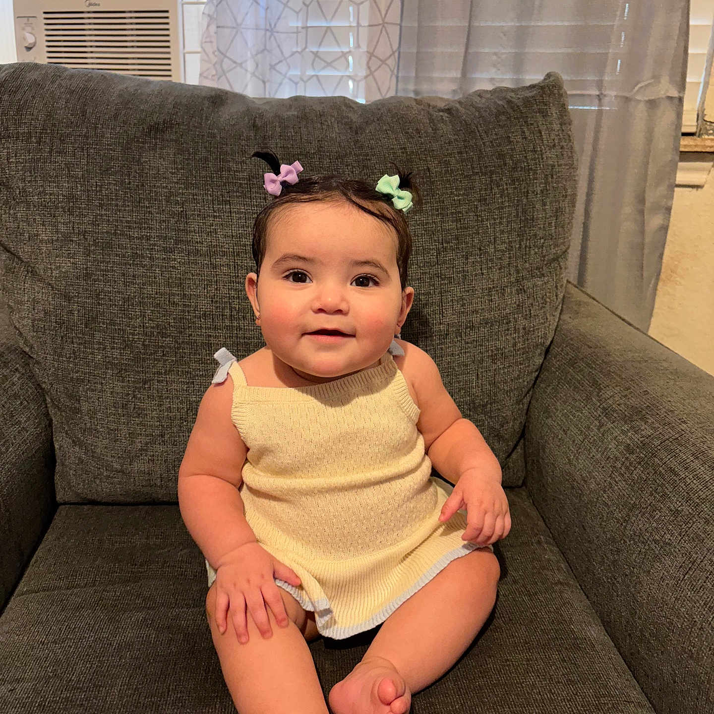 Tracey is registered to the contest to win money with this photo: baby, child, smiling, sitting, couch, gray_couch, hair_bows, indoors, window_air_conditioner, curtains, cute, portrait, infant, happy, skin, clothing, furniture, home, person, relaxed