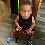 Xykie is registered to the contest to win money with this photo: toddler, child, curly_hair, indoor, wooden_box, sitting, small_stool, flooring, legs, black_shirt, blue_socks, colorful_socks, person, wood, curious, portrait, baby, hands, expression, home